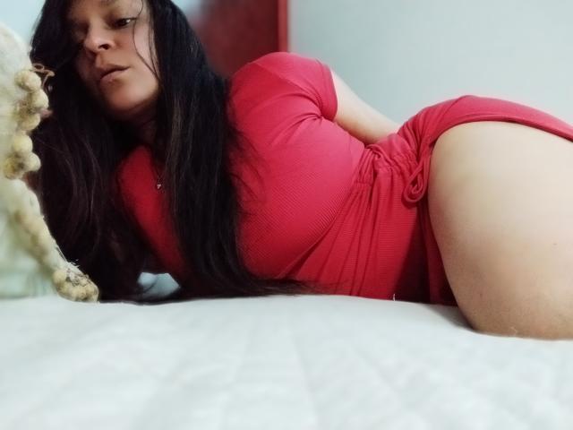 Angelonix with long black hair, red dress, lying on white bed, showing her ass, looking down, stuffed animal on left. Soft lighting, slightly blurred background.