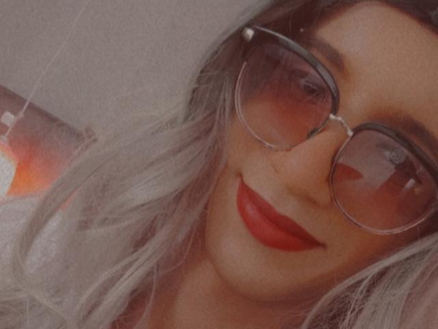 El rostro de CharlyJo, con lápiz labial rojo, cabello plateado y grandes gafas de sol redondas, sonriendo sutilmente. Los senos no son visibles, el enfoque está en el rostro y la parte superior del pecho. Iluminación suave y cálida..