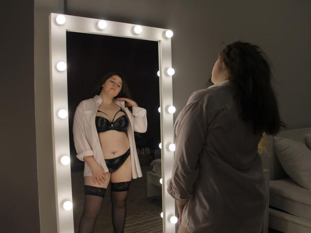 JessikaDaniels stands in front of a mirror wearing black leather bra, panties, and stockings with a white shirt, looking at her reflection. Her tits are medium, pussy is covered.