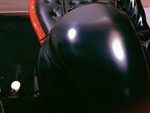 Il brillante sedere di ClaireMortin, rivestito in lattice nero, è in primo piano, riflettendo la luce, mentre sullo sfondo si trova una poltrona in pelle nera con seduta arancione..
