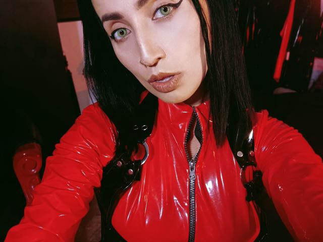 ClaireMortin, met groene ogen, draagt een glanzend rood latex topje, zwarte bandjes en opvallende eyeliner, en kijkt in de camera met een lichtjes fronsende uitdrukking, waarbij haar borsten zichtbaar zijn..
