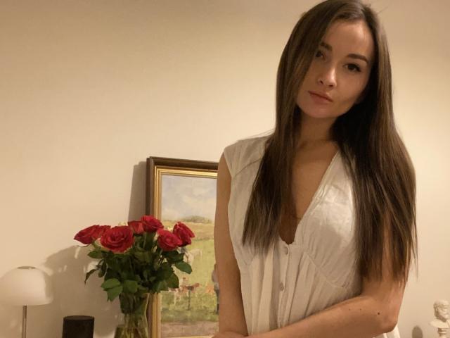 Dionisia, avec de longs cheveux bruns, une robe blanche sans manches, des roses rouges sur la table, une peinture en arrière-plan, debout, souriant légèrement, les bras croisés, regardant la caméra..