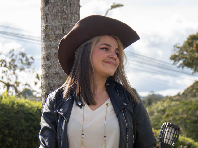 TokioParker, com cabelo castanho e loiro na altura dos ombros, usando um chapéu marrom, jaqueta de couro preta, camisa branca e colar de ouro, sorrindo com confiança em um ambiente externo ensolarado..