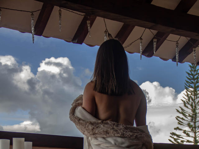 TokioParker, con la espalda desnuda y pequeños senos, con los brazos envueltos en una manta suave, debajo de un dosel de madera con cristales colgantes, mirando un cielo azul nublado..