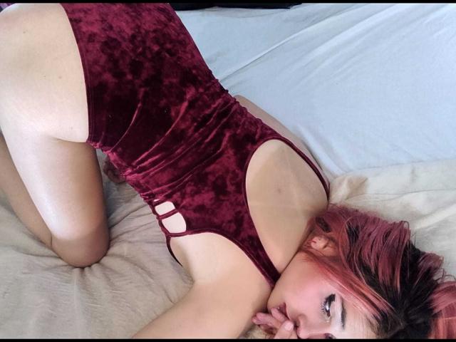 CameroonSweet est allongée sur un lit, vêtue d'une combinaison rouge en velours avec des découpes, à quatre pattes, montrant ses fesses, avec des cheveux teints en rose, et regardant la caméra..