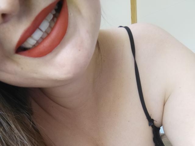 Le visage de MysteryOlympe, avec du rouge à lèvres, des dents blanches et un bandeau noir, légèrement penché, avec une partie de la poitrine visible. Peau claire, cheveux bruns, gros plan..