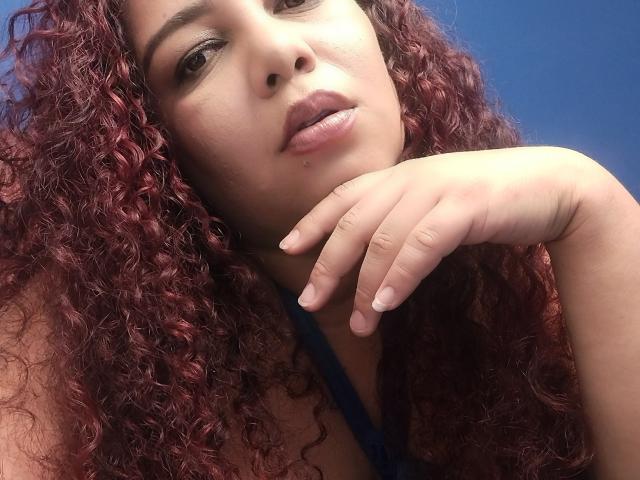 AdelineLox with curly red-purple hair, smiling, wearing a black halter top, silver necklace, and hoop earrings, has medium tits and light brown skin. Blue background.