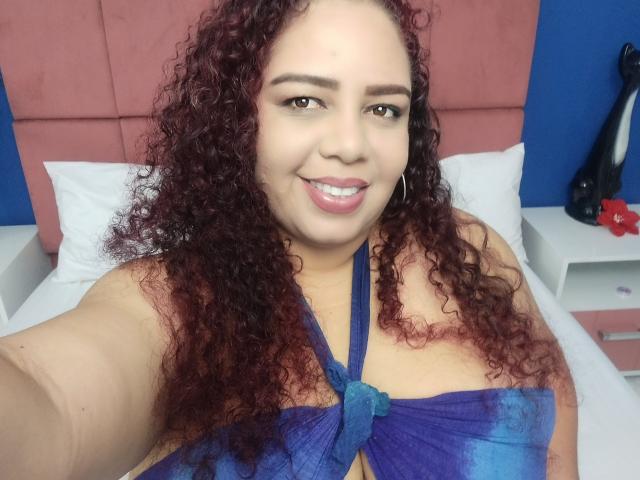 AdelineLox met krullend rood haar, roze lippen, een blauw topje met halterneck en grote borsten, kijkt naar de camera met lichtjes open mond. Op de achtergrond is een blauwe muur en een roze hoofdbord te zien..