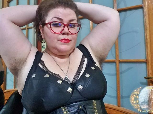 KaryQueen with big boobs in a black, studded, lace-up top, red lipstick, and a wide, black corset. Her arms are raised, showing armpit hair.