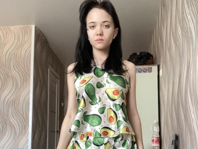 KiraLisa with black hair, wearing a sleeveless avocado print dress, standing in a room, looking straight at the camera, medium-sized tits, pale skin, and a neutral expression.