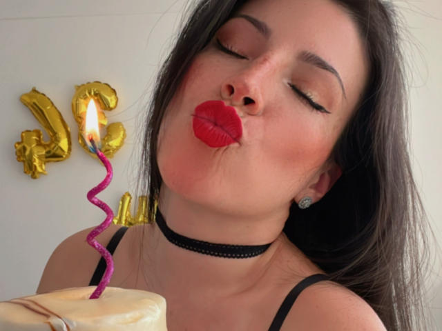 Liliysweet with her eyes closed, red lipstick, and black choker, kissing a birthday cake with a purple candle. Her tits are slightly visible, and she's wearing a black top.