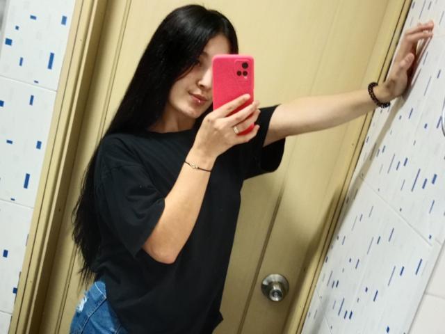 SarahGood with long black hair, wearing a black t-shirt and blue jeans, taking a mirror selfie with a pink phone, right arm against a wall.