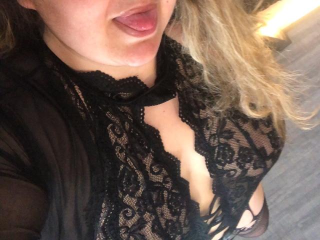 Het gezicht van AnaKelia, met haar tong uit, gekleed in zwarte kanten lingerie, waarbij haar borsten zichtbaar zijn, met golvend blond haar, en een doorschijnende zwarte badjas, op een selfie..