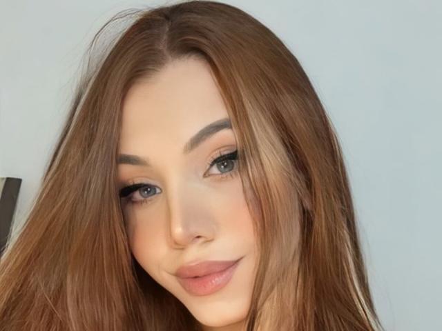 SophieFantasyHot heeft lang bruin haar, blauwe ogen en volle, roze lippen met subtiele make-up. Ze kijkt naar de camera met een lichte glimlach..