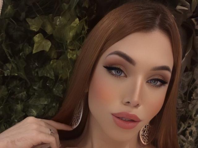 SophieFantasyHot a les cheveux bruns, les yeux bleus et porte du rouge à lèvres rose, avec du crayon noir pour les yeux et des boucles d'oreilles en dentelle. Elle a une main près de son visage et regarde légèrement sur le côté..