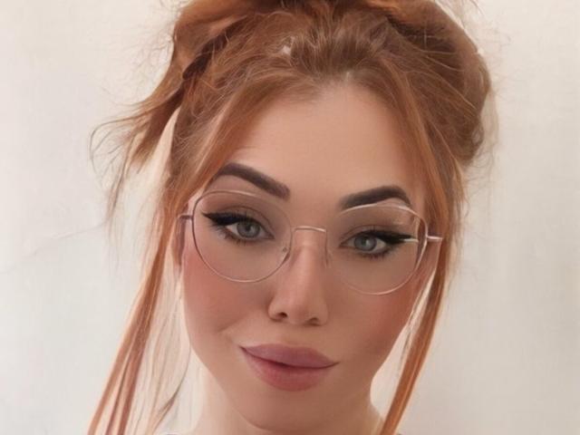 A SophieFantasyHot tem cabelo ruivo preso em um coque bagunçado, usa óculos redondos, tem olhos azuis e está sorrindo com batom rosa. Seus seios não estão visíveis..