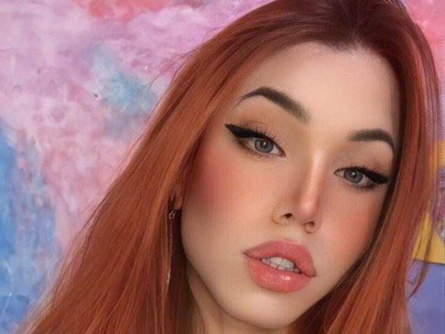SophieFantasyHot has red hair, blue eyes, thick eyebrows, black eyeliner, pink lips, and a cute face with a slight pout. Her skin is fair and her makeup is on point.