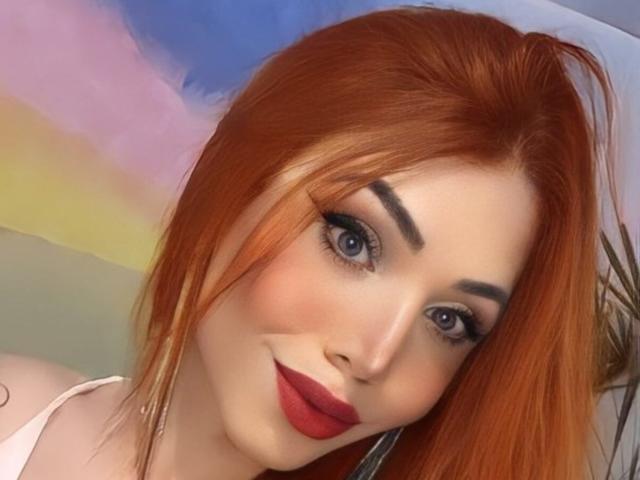 Sophie, une femme séduisante aux cheveux roux, aux yeux bleus et au rouge à lèvres rouge, portant de l'eye-liner noir, souriant à la caméra et mettant en valeur ses jolies fesses. Ses sourcils sont parfaitement dessinés..