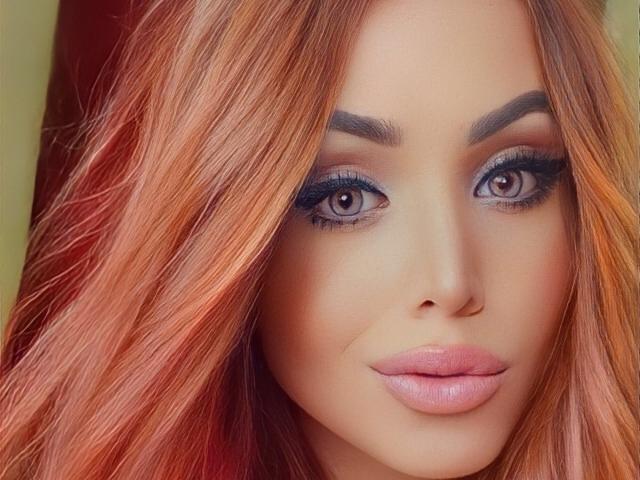 SophieFantasyHot hat rot-orangefarbenes Haar, volle, rosafarbene Lippen und grau-blaue Augen mit dunklem Eyeliner. Ihre Augenbrauen sind dick und gebogen, und sie schaut direkt in die Kamera..