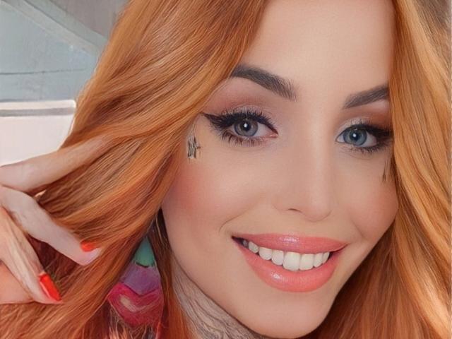 SophieFantasyHot tiene cabello rojo, ojos azules y una sonrisa con lápiz labial rosa. Tiene delineador de ojos negro, un tatuaje pequeño debajo del ojo derecho y uñas rojas..