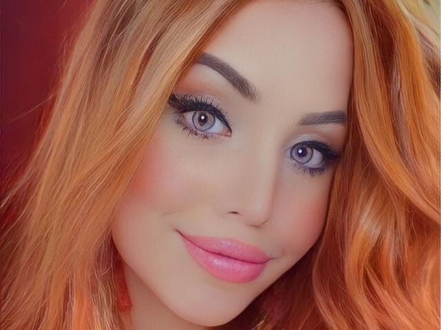 A SophieFantasyHot tem cabelo laranja, olhos azuis, sobrancelhas grossas, batom rosa e um sorriso adorável. Sua maquiagem inclui delineador escuro e cílios longos. Ela parece sexy..