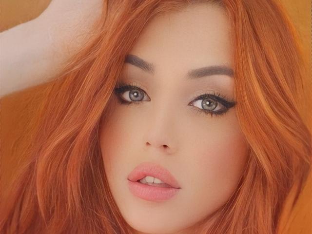 SophieFantasyHot tiene cabello rojo, ojos verdes y labios rosados con una apariencia ligeramente entreabierta y sensual. Su mano está en su cabello, resaltando su maquillaje y su atractivo rostro..