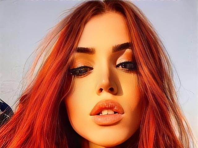 SophieFantasyHot tiene cabello rojo, labios carnosos y ojos azules con delineador de ojos oscuro. Su rostro atractivo está centrado, con la boca ligeramente abierta, mostrando sus dientes superiores..