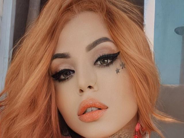 SophieFantasyHot has orange hair, thick eyebrows, black winged eyeliner, orange lipstick, and a star tattoo near her right eye. Her lips are slightly parted.