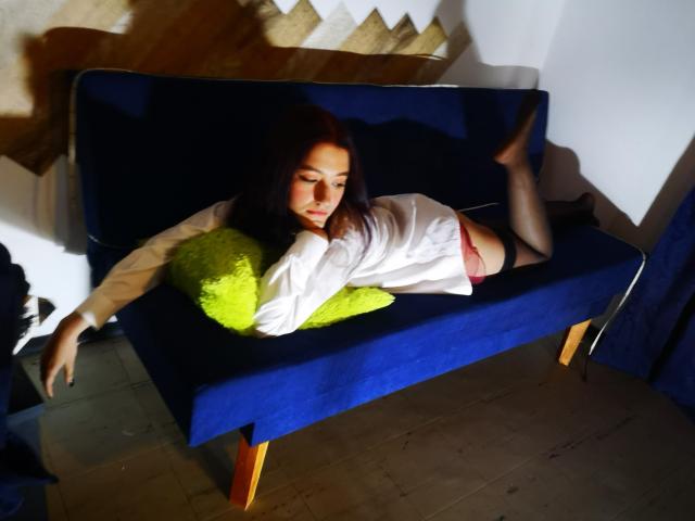 RukkiaParist, met lang donker haar, ligt op een blauwe fluwelen bank, gekleed in een wit shirt, rode onderbroek, zwarte kousen, en met een groen kussen, met de benen omhoog en de ogen gesloten..
