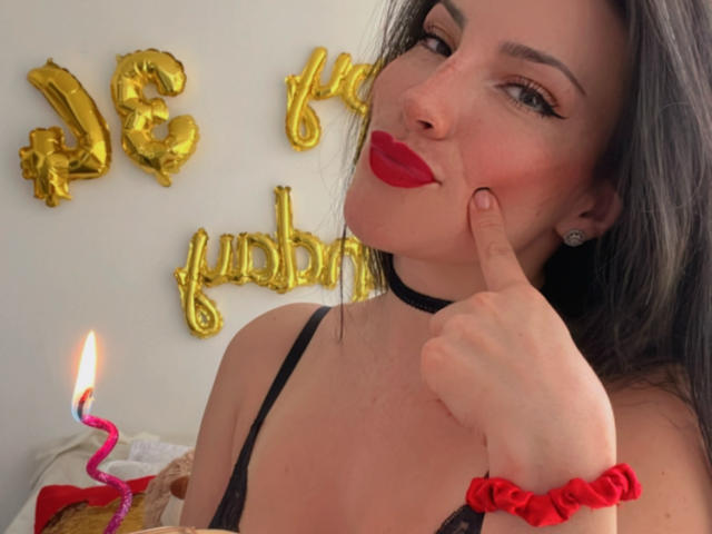 Liliysweet with red lipstick, black choker, and red scrunchie, pointing at her cheek, black lace bra, candle, and gold balloons. Her tits are small, and she's cute.