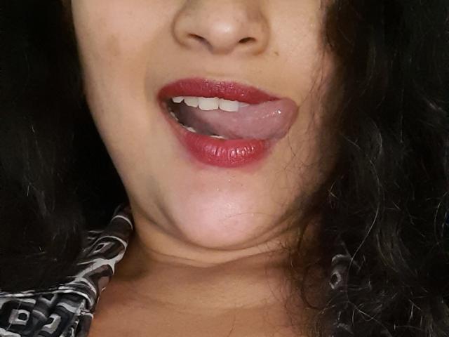 Xuuley's face, tongue out, red lipstick, slightly open mouth, showing teeth, black wavy hair, light skin, wearing a patterned shirt, visible collarbone.