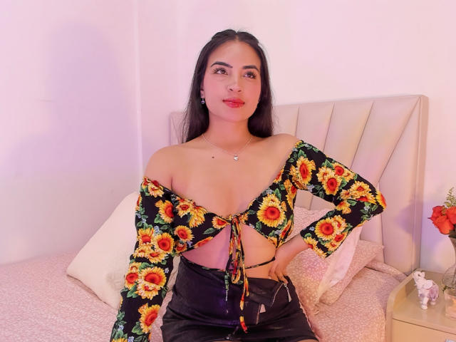 Emily Jensenn, con una blusa de girasol que deja ver sus pechos, pantalones de cuero negro, cabello largo y negro, lápiz labial rojo y aretes verdes, sentada en una cama con un cabecero blanco..