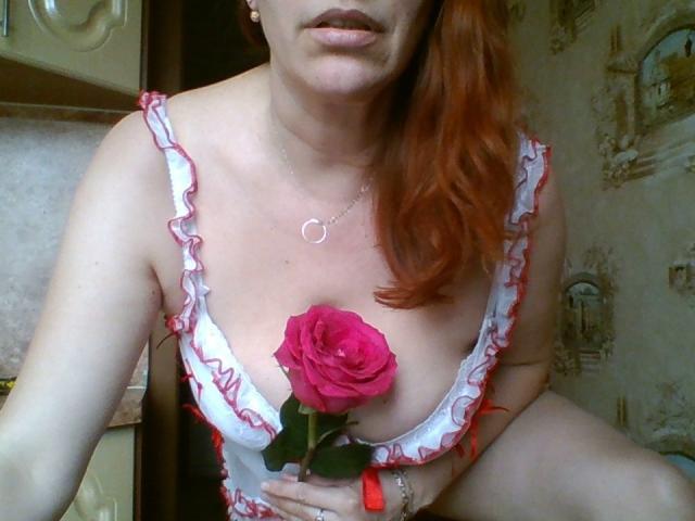 MissieLorrie with red hair, holding a pink rose between her tits, wearing a white dress with red ruffles, and a silver necklace, in a cozy indoor setting.