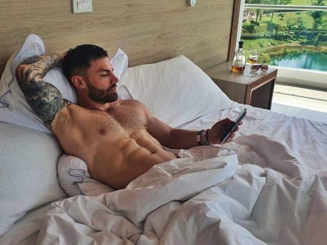 Ryzen, shirtless with a tattooed arm, lies in bed with white sheets, holding a phone, his muscular, hairy chest exposed, a glass of whiskey on the wooden nightstand.