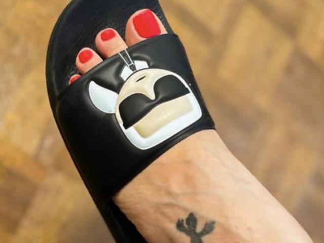 SiPassioneX's foot with red-painted toenails wearing a black sandal featuring a goofy, white-and-black face buckle, showcasing a small black tattoo on the ankle.
