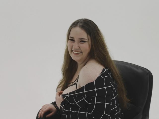 DaniellaGraceZZ, with long brown hair, smiling, pulling down a black and white checkered shirt to reveal her small tits, sitting in a black chair, looking cute.