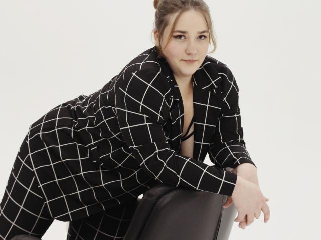 DaniellaGraceZZ with brown hair in a bun, wearing a black checkered coat, leaning on a chair, showing her tits in a black strappy bra, looking at the camera.