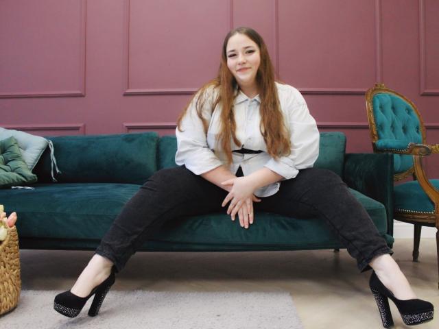 DaniellaGraceZZ sits on a green velvet sofa with legs spread, wearing a white shirt, black pants, and black high heels. Her hands rest on her pussy.