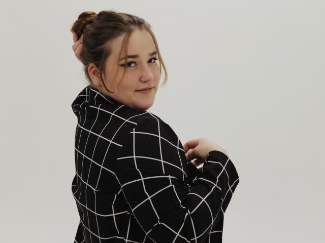 DaniellaGraceZZ with brown hair in a bun, wearing a black and white checkered hoodie, looking back with a slight smile, hands near her chest. Soft, cute, sexy.