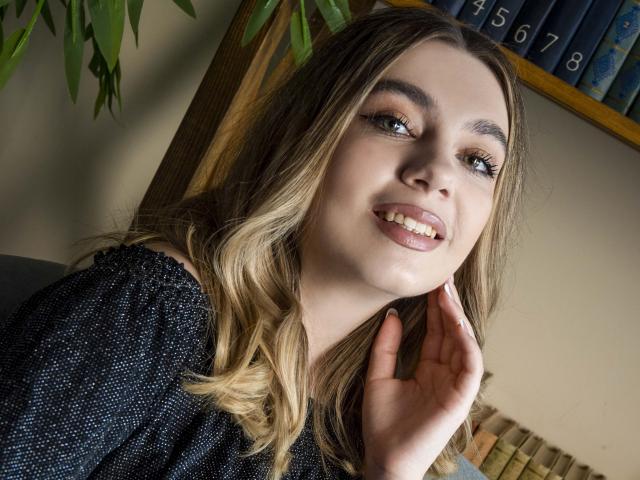 MilanaKitty a les cheveux blonds ondulés, une peau claire, et elle sourit avec la langue légèrement sortie, portant un haut noir à pois, sa main près de son visage..