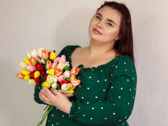 KaidoKun avec des cheveux roux, une robe à pois verts, tenant un bouquet coloré de tulipes jaunes, roses et blanches, la tête légèrement inclinée, un sourire doux, des yeux verts, et une bague à la main gauche..