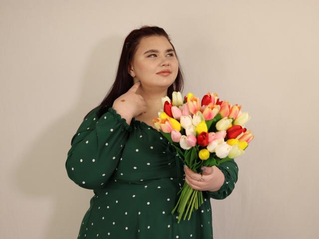 KaidoKun mit dunklem Haar, in einem grünen, gepunkteten Kleid, hält einen bunten Strauß Tulpen, zeigt Daumen hoch, lächelt leicht und steht vor einer schlichten Wand..