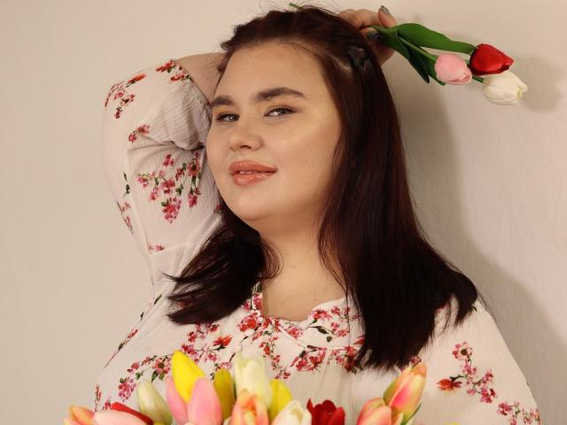KaidoKun met donkerbruin haar, die roze en witte tulpen in één hand houdt, een witte blouse met bloemen aanheeft, glimlacht met licht open roze lippen en naar de zijkant kijkt..
