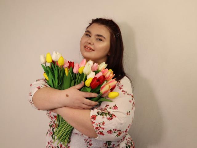 KaidoKun com pele clara, cabelo castanho, vestido floral, segurando um buquê de tulipas amarelas, rosa e brancas, sorrindo, braços cruzados, com uma pinta visível na mão esquerda..