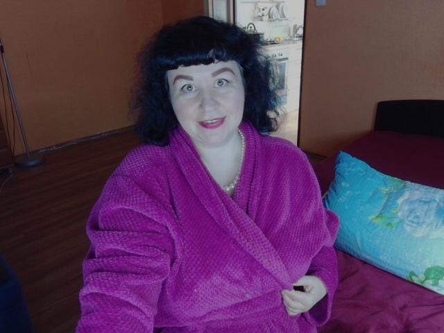 ErinSmart, con cabello negro rizado, piel clara y lápiz labial rosa, vestida con una bata morada de textura waffle, sentada en una cama con un cojín azul y una manta roja..