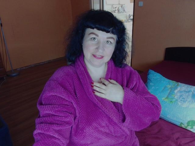 Erin, con i capelli neri ricci, la pelle chiara e il rossetto rosso, indossa un accappatoio viola e morbido, tiene una mano sul petto, sorride e mostra le unghie dipinte di rosso..