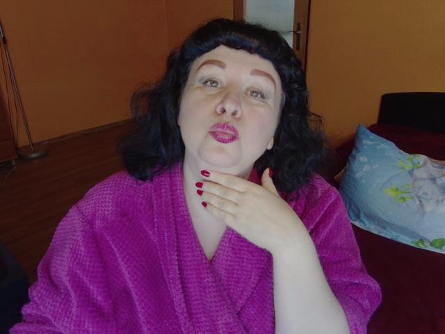 ErinSmart with curly black hair, pink lipstick, and red nails, wearing a purple bathrobe, has her hand on her neck, pouting slightly. Her tits are visible under the robe.