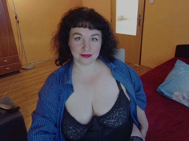 ErinSmart mit lockigem, schwarzem Haar, blau kariertem Hemd, offen, schwarzer Spitzen-BH, der große Brüste zeigt, rotem Lippenstift, sitzend auf einem Bett mit einer roten Decke, Holzboden und Holztür..