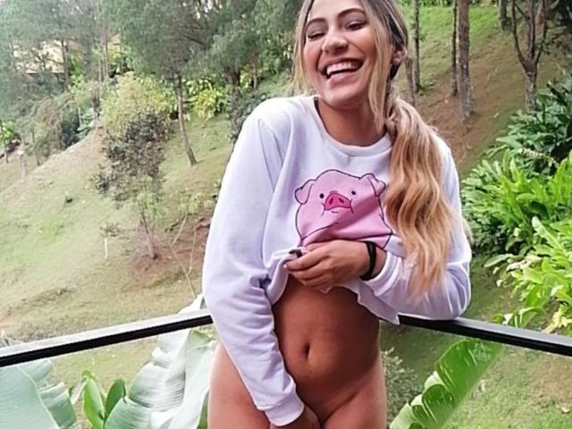KataMillerr with blonde ponytail, smiling, lifting white sweatshirt showing her bare, tan-skinned, flat-stomached pussy, wearing pink pig graphic, standing on green, leafy balcony.