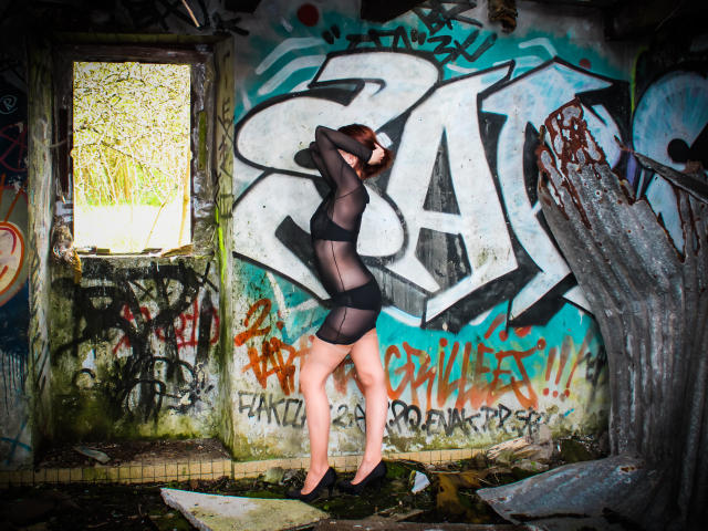 BKLove está de pie, descalza, en una habitación llena de grafitis, vistiendo un vestido de malla negra transparente, con los brazos levantados, mostrando su trasero y sus pequeños senos. Sus piernas son delgadas y está descalza..